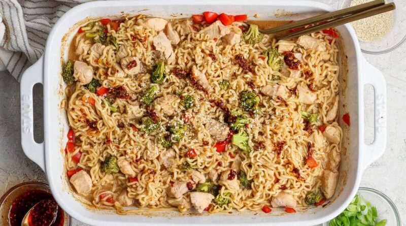 Rectangular white baking dish filled with cooked ramen noodles, chicken pieces, broccoli, and red peppers, garnished with sesame seeds and sauce, with chopsticks resting on the dish.