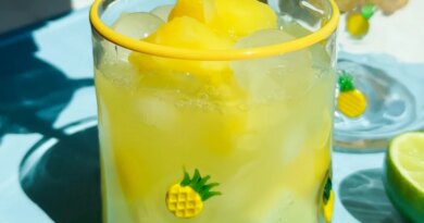 A glass decorated with small pineapples is filled with yellow frozen drink and ice, with juice being poured in. Lime slices are on the blue surface beside the glass.