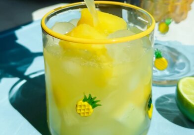 A glass decorated with small pineapples is filled with yellow frozen drink and ice, with juice being poured in. Lime slices are on the blue surface beside the glass.
