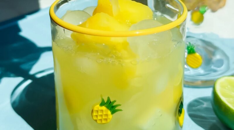 A glass decorated with small pineapples is filled with yellow frozen drink and ice, with juice being poured in. Lime slices are on the blue surface beside the glass.