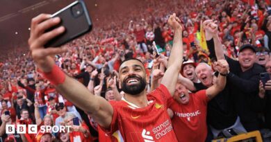A grinning Mohamed Salah, in red club kit, takes a selfie with a mobile phone in front of excited Liverpool supporters