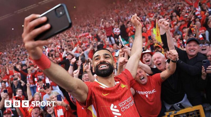 A grinning Mohamed Salah, in red club kit, takes a selfie with a mobile phone in front of excited Liverpool supporters