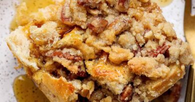 A close-up of a slice of pecan streusel bread pudding topped with pecans and syrup, served on a speckled plate with a fork.