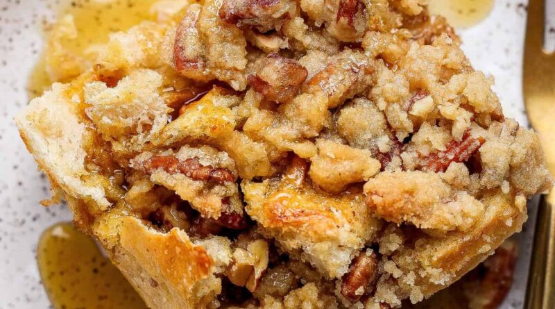 A close-up of a slice of pecan streusel bread pudding topped with pecans and syrup, served on a speckled plate with a fork.