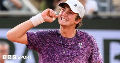 Joao Fonseca, wearing a purple top with white flecks and a white cap, grins and puts his right hand behind his ear to appreciate the support of the crowd