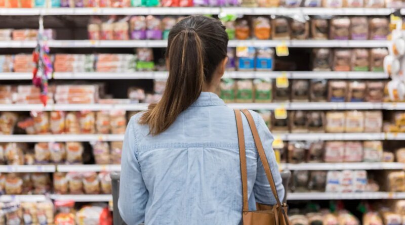 In this rear view, an unrecognizable woman stands with a shopping cart in front of a shelf full of food in the bread aisle of a grocery store.