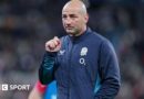 Steve Borthwick, wearing a nany blue zip-up England jacket, looks into the distance, his right hand raised to shoulder level