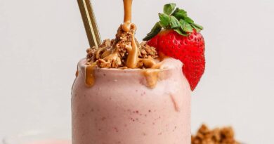 A strawberry smoothie topped with granola, peanut butter, a fresh strawberry, and two straws in a glass.