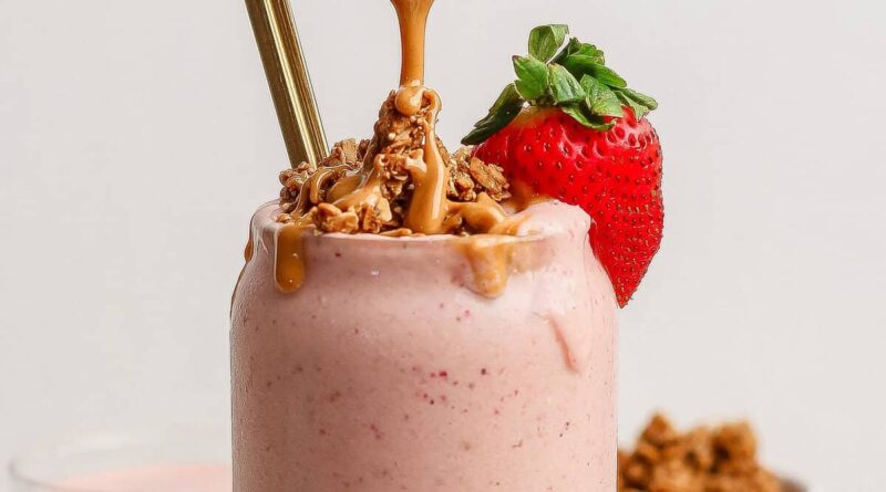 A strawberry smoothie topped with granola, peanut butter, a fresh strawberry, and two straws in a glass.