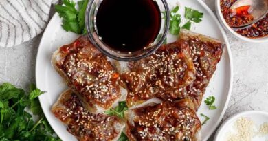 A white plate with five pieces of crispy, glazed tofu topped with sesame seeds, garnished with cilantro, and served with a small bowl of dark dipping sauce.