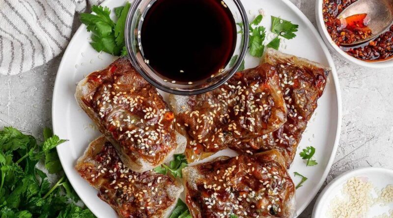 A white plate with five pieces of crispy, glazed tofu topped with sesame seeds, garnished with cilantro, and served with a small bowl of dark dipping sauce.