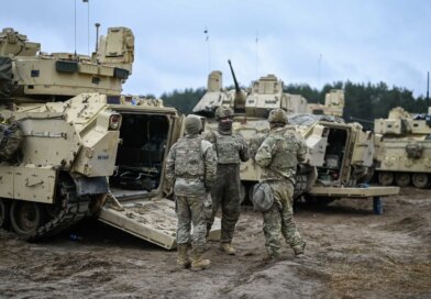 Soldiers in camouflage near the open rear of a pale armored vehicle standing on mud.