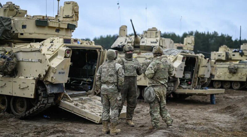 Soldiers in camouflage near the open rear of a pale armored vehicle standing on mud.