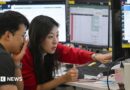 A woman in a red cardigan points to a screen during a discussion with a colleague in a trading room in Seoul 