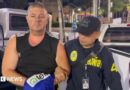 Simon Peter Carman, a man with short light brown hair wearing a sleeveless black too, is escorted by a Thai police officer.
