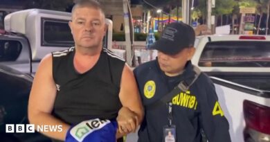 Simon Peter Carman, a man with short light brown hair wearing a sleeveless black too, is escorted by a Thai police officer.