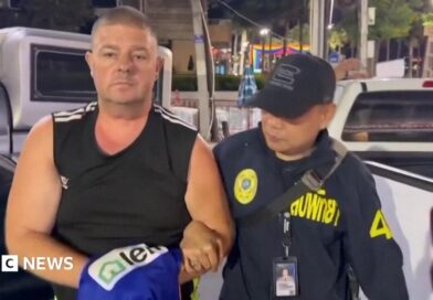 Simon Peter Carman, a man with short light brown hair wearing a sleeveless black too, is escorted by a Thai police officer.