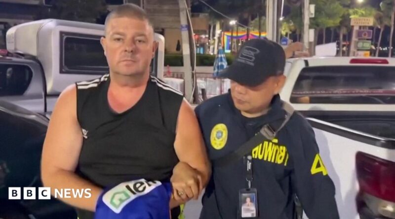 Simon Peter Carman, a man with short light brown hair wearing a sleeveless black too, is escorted by a Thai police officer.