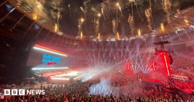 A huge crowd fills the Tottenham Hotspur Stadium, illuminated by stage lights. Fireworks go off from the roof. A stage is lit brightly. Behind, the big screen says Debí Tirar Más Fotos World Tour.   