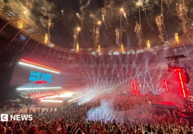 A huge crowd fills the Tottenham Hotspur Stadium, illuminated by stage lights. Fireworks go off from the roof. A stage is lit brightly. Behind, the big screen says Debí Tirar Más Fotos World Tour.   