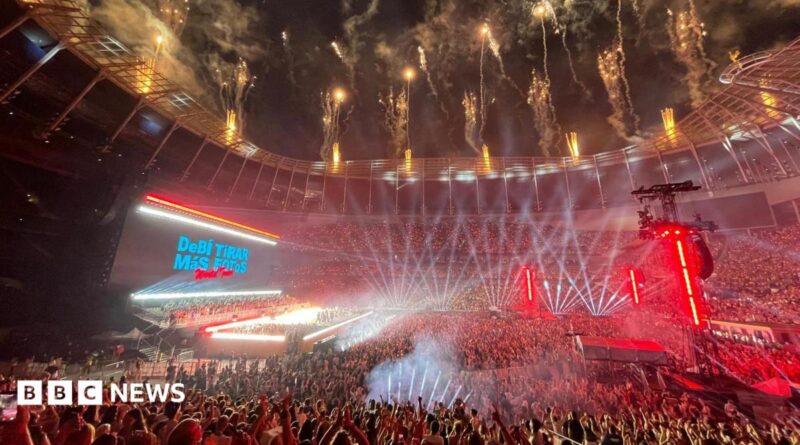 A huge crowd fills the Tottenham Hotspur Stadium, illuminated by stage lights. Fireworks go off from the roof. A stage is lit brightly. Behind, the big screen says Debí Tirar Más Fotos World Tour.
