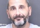 A custody shot of Matthew Ashley Foster-Smith. He has short hair and a beard and is wearing a grey jumper. 