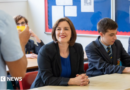 Phillipson wears a black suit jacket over a royal blue dress with a silver necklace. Her hair is worn in a dark brown, chin-length bob. She wears bright red lipstick and is smiling at a teacher, who can partially be seen in the foreground on the left. There are pupils sitting around her, all wearing a navy school uniform with a pastel blue shirt and a navy tie with pale blue and gold diagonal stripes. 