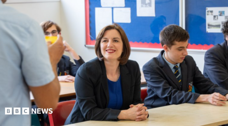 Phillipson wears a black suit jacket over a royal blue dress with a silver necklace. Her hair is worn in a dark brown, chin-length bob. She wears bright red lipstick and is smiling at a teacher, who can partially be seen in the foreground on the left. There are pupils sitting around her, all wearing a navy school uniform with a pastel blue shirt and a navy tie with pale blue and gold diagonal stripes.