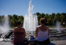 Two women look at a fountain on a sunny day with their backs to the camera