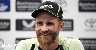Kane Williamson smiling and wearing a black New Zealand cap sits in front of two long black microphones