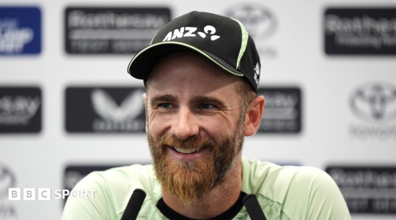 Kane Williamson smiling and wearing a black New Zealand cap sits in front of two long black microphones