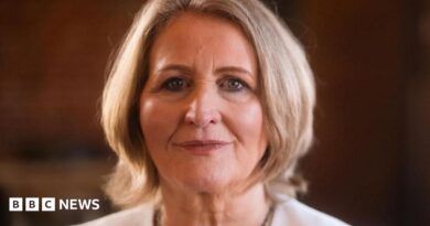 A woman with short blonde hair wearing a white suit jacket smiles faintly at the camera