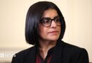 Home Secretary Shabana Mahmood is pictured looking off camera. She is wearing a dark jacket with a red and black top on beneath. 