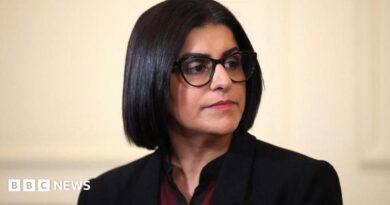 Home Secretary Shabana Mahmood is pictured looking off camera. She is wearing a dark jacket with a red and black top on beneath. 
