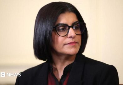 Home Secretary Shabana Mahmood is pictured looking off camera. She is wearing a dark jacket with a red and black top on beneath. 