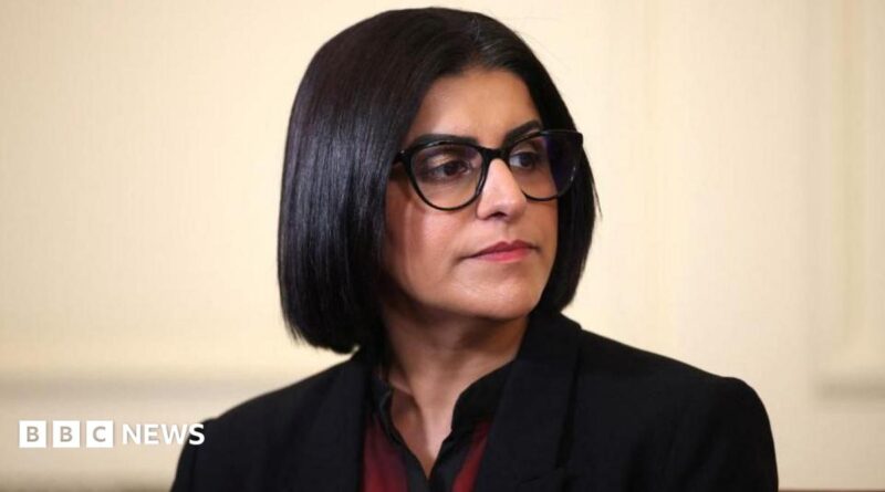 Home Secretary Shabana Mahmood is pictured looking off camera. She is wearing a dark jacket with a red and black top on beneath.