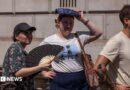 A woman fans herself as another shelters from the sun as they wait to take a picture in front of a phone box in Parliament Square. In front of them is a man wearing a beige top.