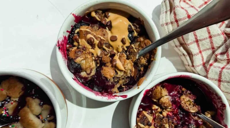 Three small white ramekins filled with baked berry dessert, topped with peanut butter and chocolate chips, with metal spoons and a red-striped cloth nearby.