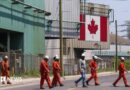 A Canadian national flag is seen in the background as seven workers dressed in orange work suits and white hard hats cross the street in front of ArcelorMittal Dofasco's steel manufacturing buildings in Hamilton, Ontario. 