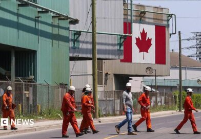 A Canadian national flag is seen in the background as seven workers dressed in orange work suits and white hard hats cross the street in front of ArcelorMittal Dofasco's steel manufacturing buildings in Hamilton, Ontario. 