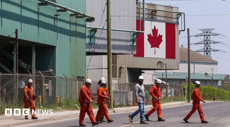 A Canadian national flag is seen in the background as seven workers dressed in orange work suits and white hard hats cross the street in front of ArcelorMittal Dofasco's steel manufacturing buildings in Hamilton, Ontario.