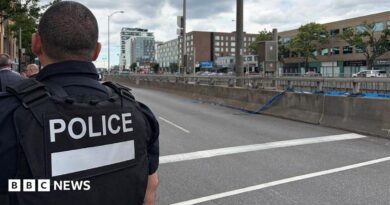 Back of officer in black vest marked with white letters reading "POLICE" next to an empty roadway with buildings across the street