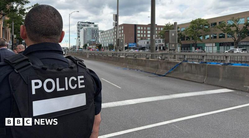 Back of officer in black vest marked with white letters reading "POLICE" next to an empty roadway with buildings across the street
