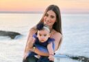 A woman with long dark hair smiles as she holds a toddler in a dress on a beach, the sunset behind her