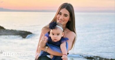 A woman with long dark hair smiles as she holds a toddler in a dress on a beach, the sunset behind her