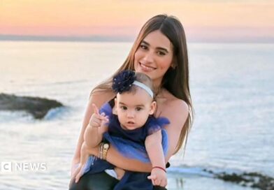 A woman with long dark hair smiles as she holds a toddler in a dress on a beach, the sunset behind her