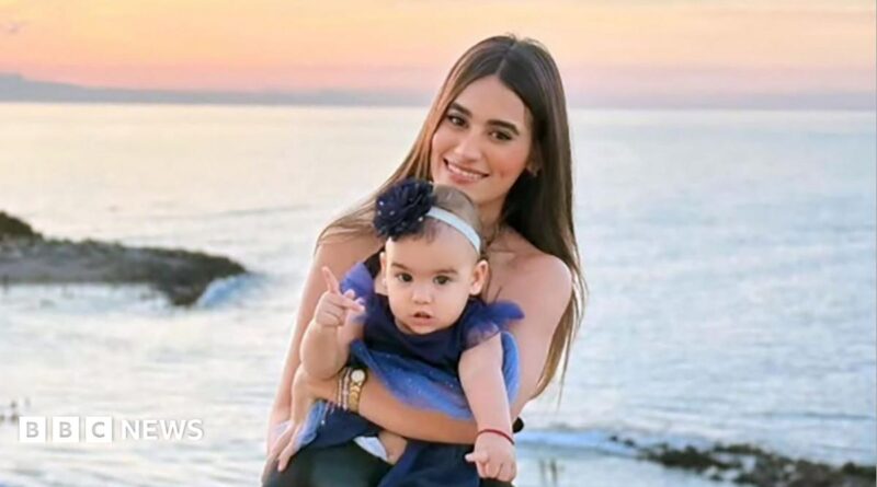 A woman with long dark hair smiles as she holds a toddler in a dress on a beach, the sunset behind her