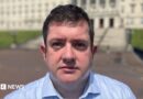 Dr David Farren, a man with dark hair, wearing a blue shirt, standing in front of Stormont Parlaiment Buildings.