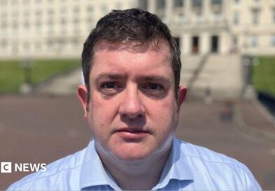 Dr David Farren, a man with dark hair, wearing a blue shirt, standing in front of Stormont Parlaiment Buildings.