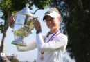 Nelly Korda, wearing a long-sleeved white top and white visor and with a gold medal on a red, white and blue ribbon around her neck, smiles and poses with the silver trophy, holding it up to her right